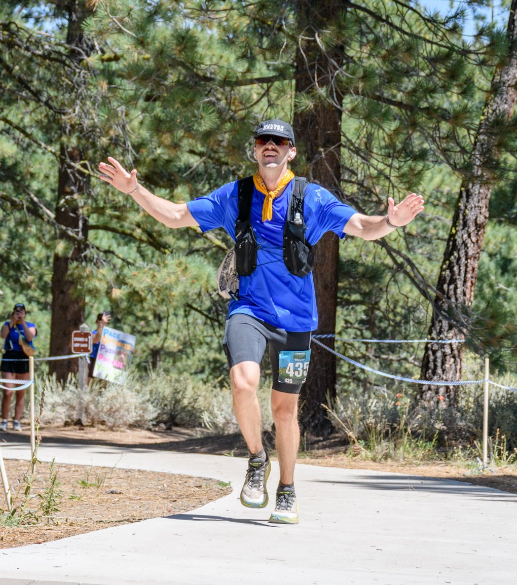 Matt finishing the Marlette 50K on a paved trail through Sierra pines, arms outstretched and grinning, wearing a blue shirt, yellow bandana, and hydration vest, race bib 435.