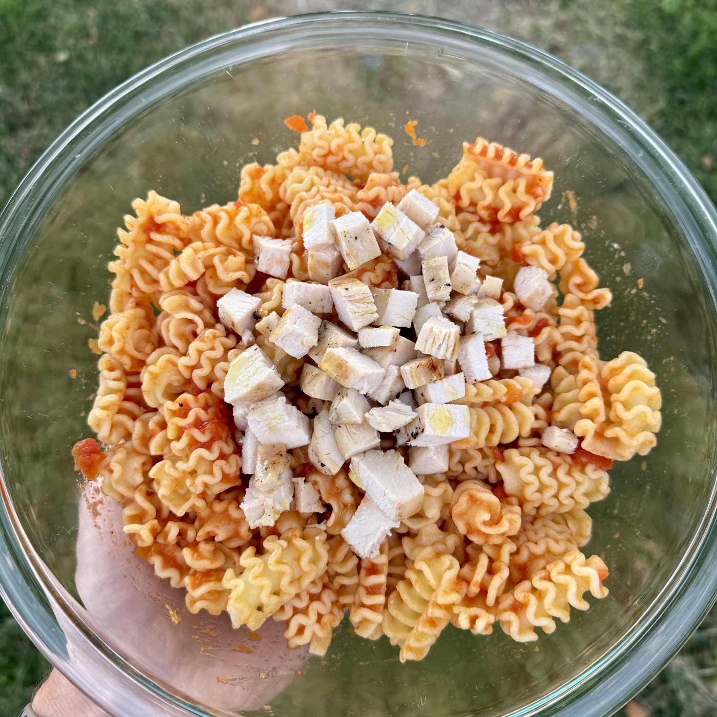 Loading-day dinner: a glass bowl of chicken marinara pasta held outdoors, radiatore pasta coated in tomato sauce with cubed grilled chicken on top.